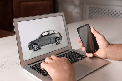 Photo of Man using laptop and phone to buy car at wooden table indoors, closeup