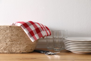 Kitchen towel in wicker basket and clean dishware on wooden table near white wall