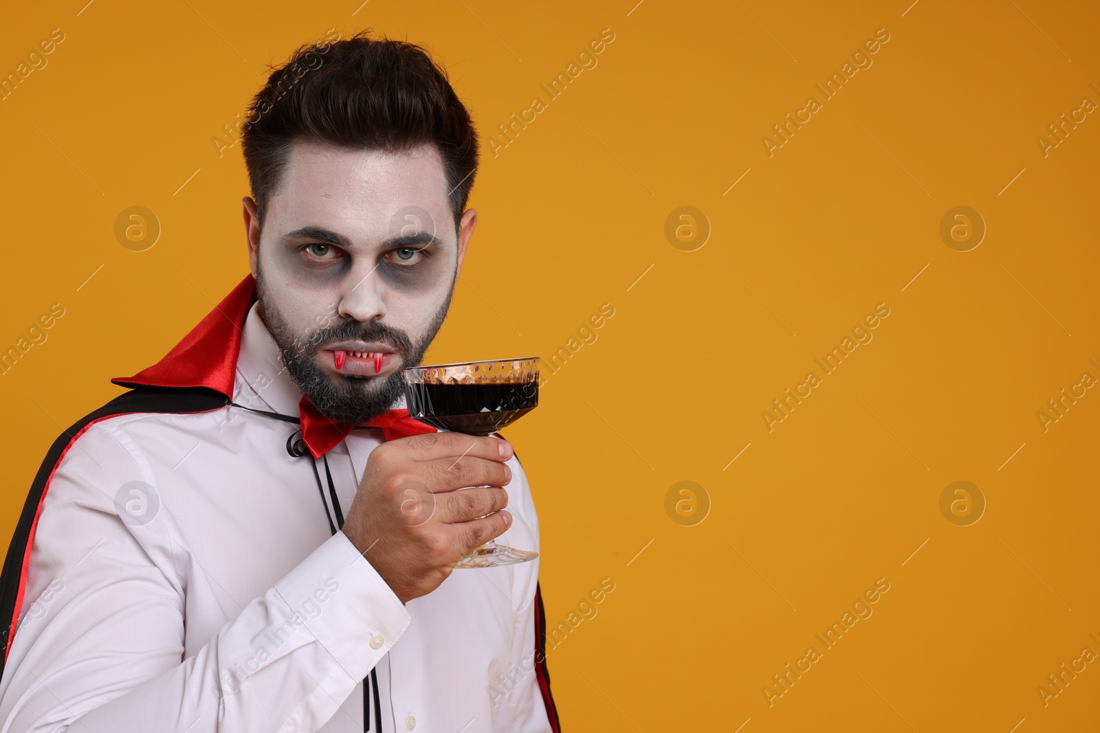 Photo of Man in scary vampire costume with fangs and glass of wine on orange background, space for text. Halloween celebration