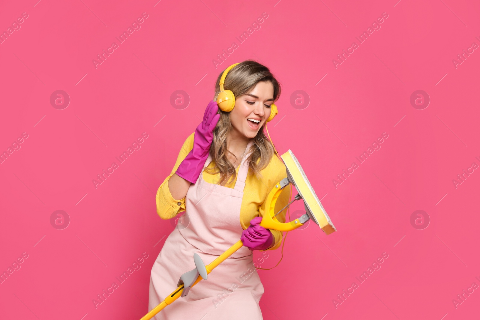 Photo of Beautiful young woman with headphones and mop singing on pink background