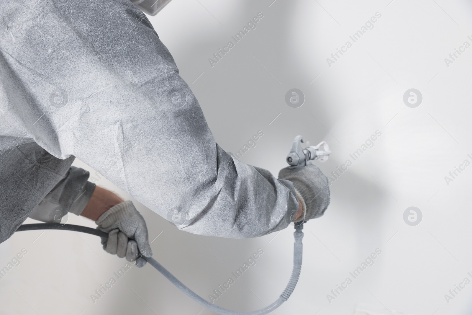 Photo of Decorator in protective overalls painting wall with spray gun indoors, closeup