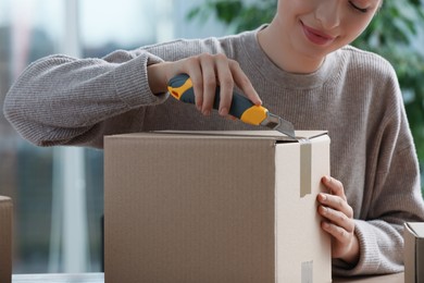 Young woman using utility knife to open parcel indoors, closeup