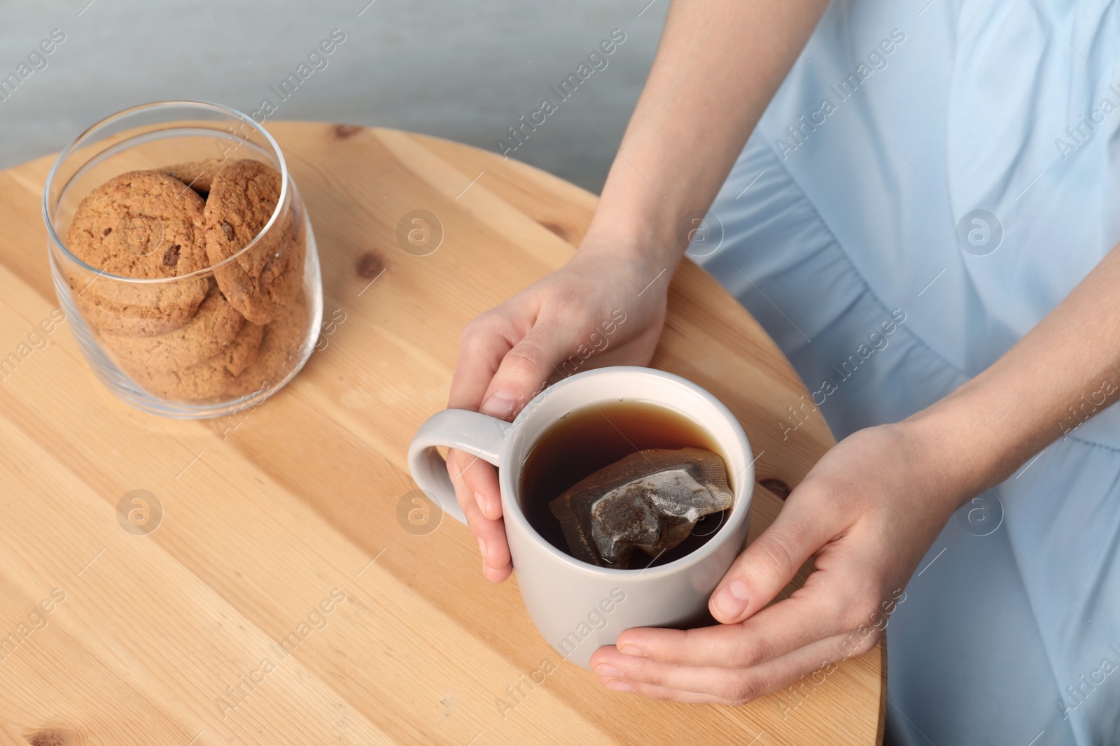 Photo of Woman holding cup of hot tea near jar with cookies at wooden table, closeup