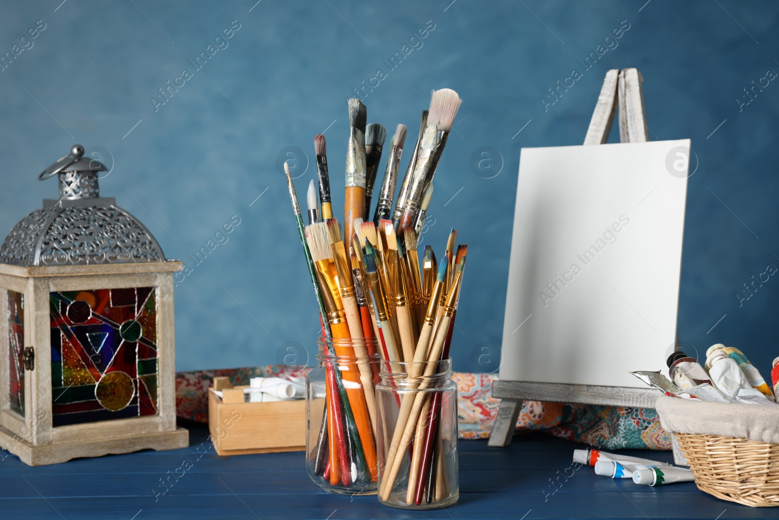 Photo of Many brushes, easel with blank canvas and paints on blue wooden table