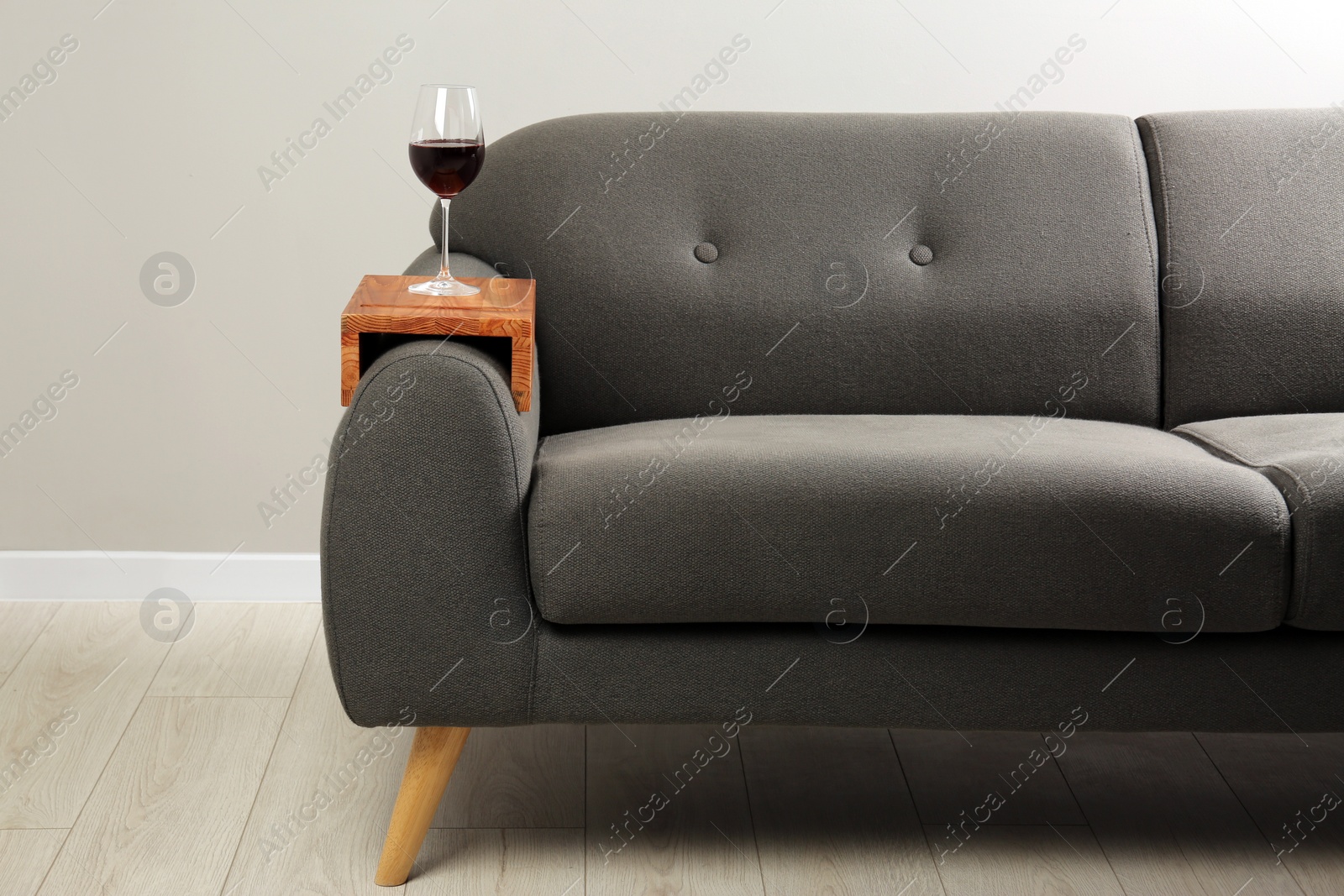 Photo of Glass of red wine on sofa with wooden armrest table in room. Interior element