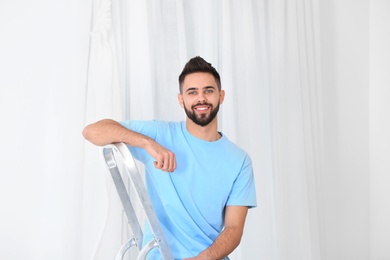 Young man sitting on stepladder near window with curtain in room