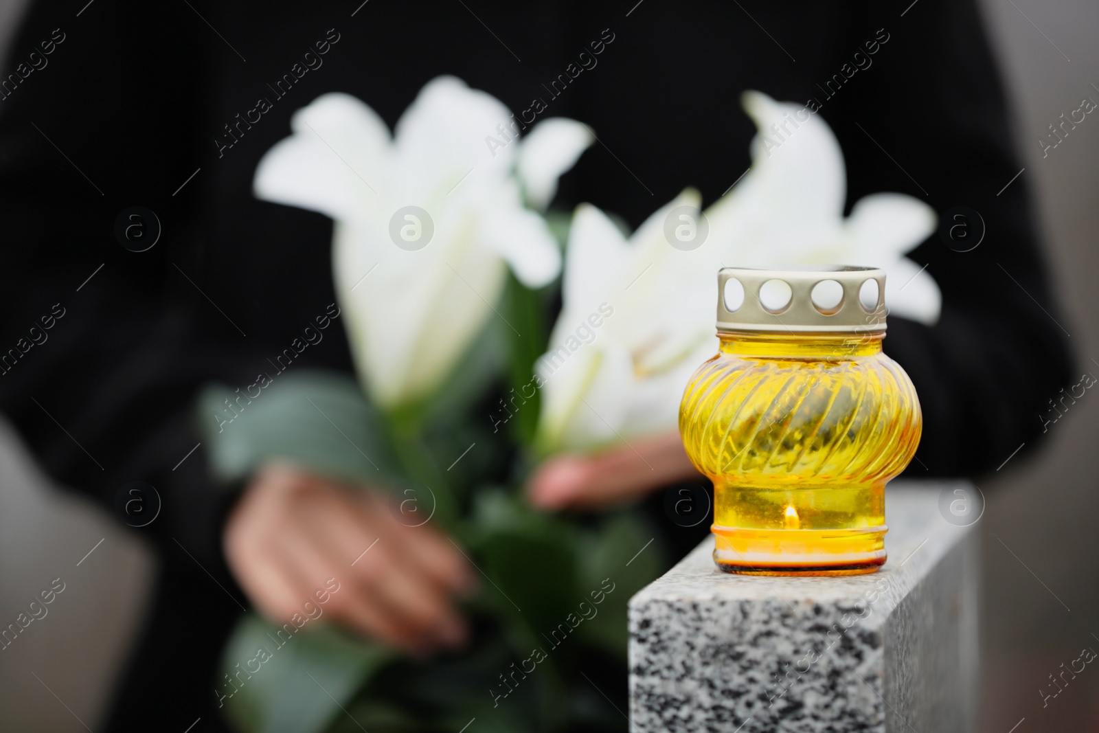 Photo of Woman holding white lilies near grey granite tombstone outdoors, focus on candle. Funeral ceremony