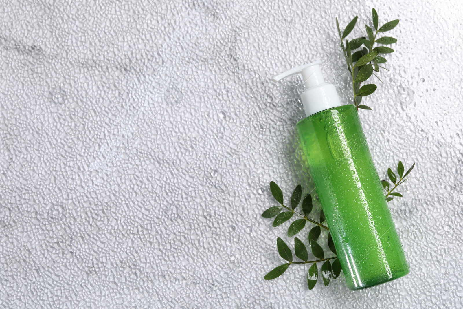 Photo of Bottle of face cleansing product and green leaves on white textured table, flat lay. Space for text