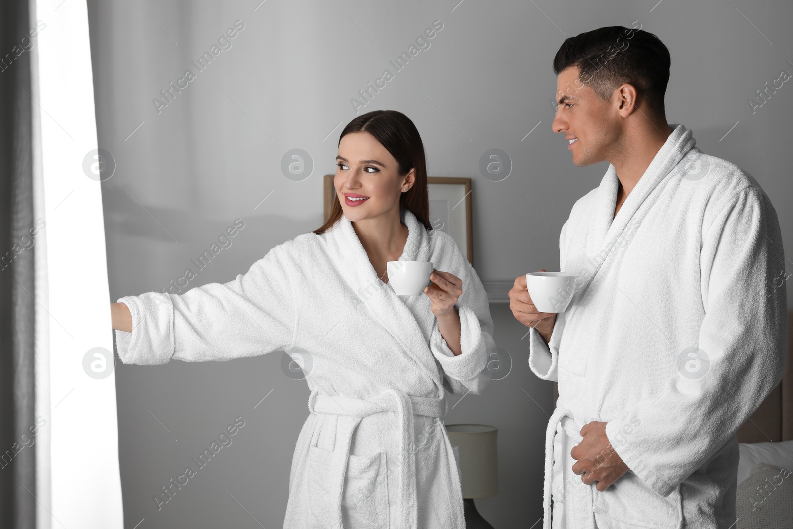 Photo of Happy couple wearing bathrobes near window in room