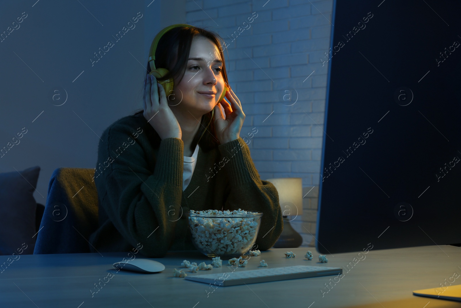 Photo of Beautiful young woman in headphones with popcorn watching film on computer at table indoors at night