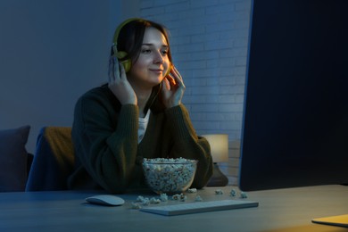 Photo of Beautiful young woman in headphones with popcorn watching film on computer at table indoors at night