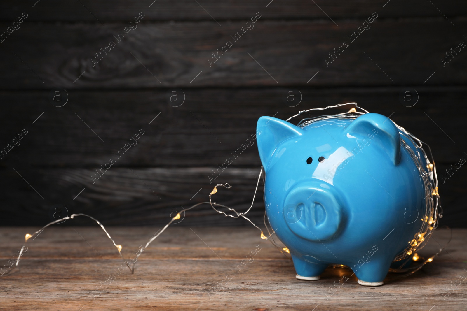 Photo of Piggy bank with fairy lights on wooden table. Space for text