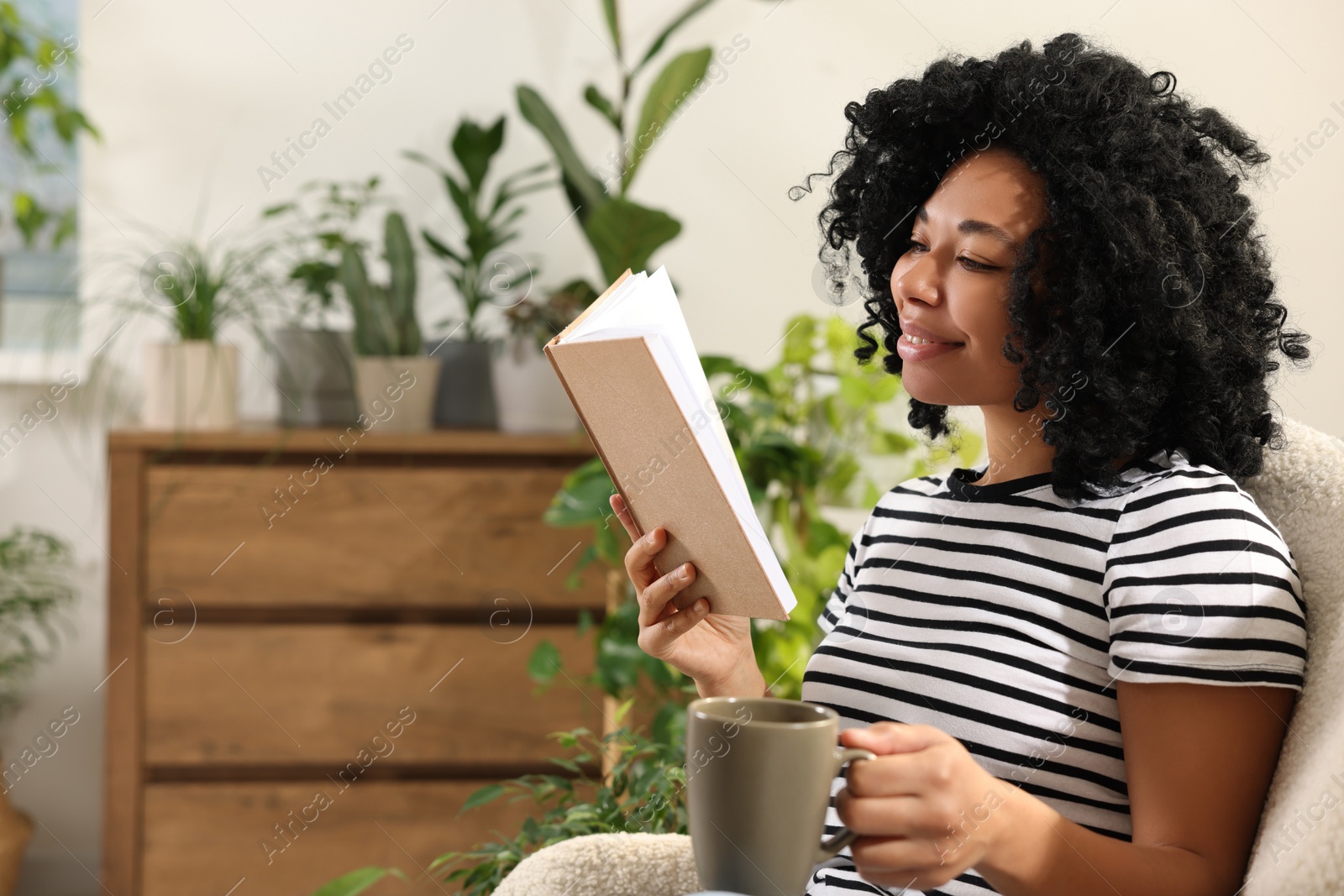 Photo of Relaxing atmosphere. Happy woman with cup of hot drink reading book near houseplants at home. Space for text