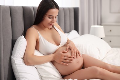 Beautiful pregnant woman in stylish comfortable underwear making heart with hands on her belly at home