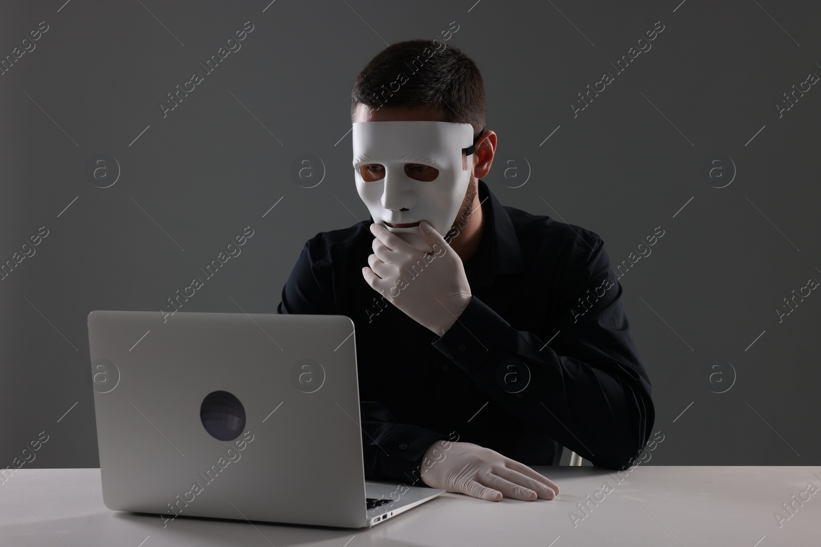 Photo of Man in mask and gloves sitting with laptop at white table against grey background