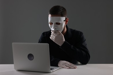 Man in mask and gloves sitting with laptop at white table against grey background