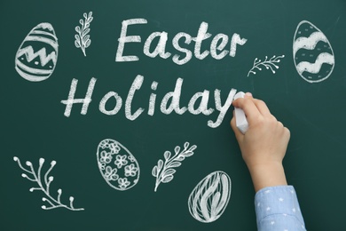 Image of Little child writing phrase Easter Holiday on green chalkboard with drawings, closeup. School break