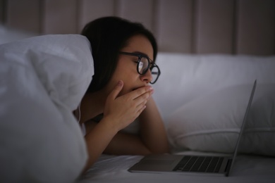 Young woman with nomophobia yawning while using laptop in bed at night. Insomnia concept