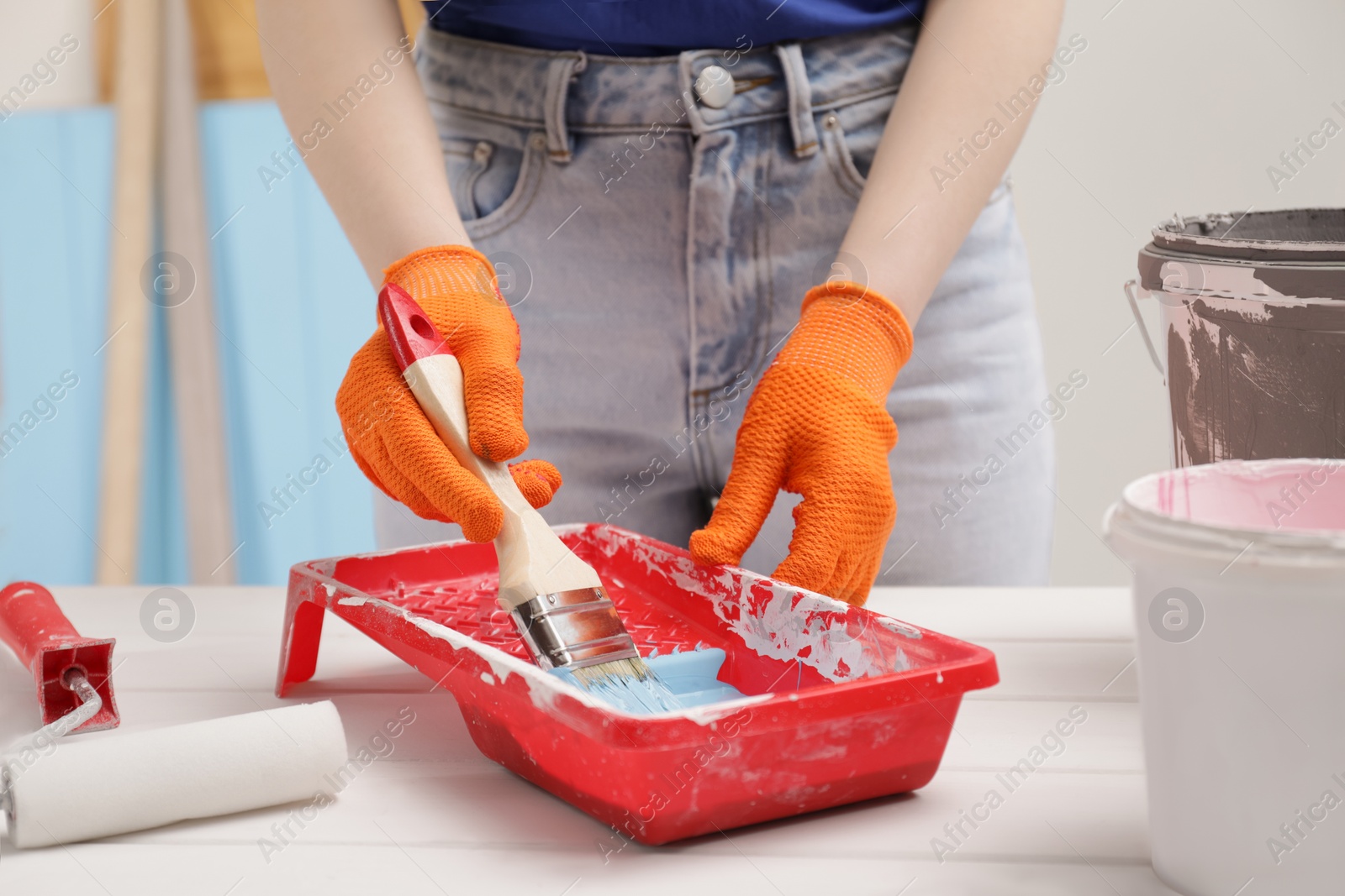 Photo of Woman taking light blue paint with brush from tray at white wooden table indoors, closeup