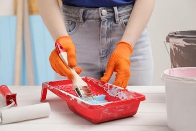 Woman taking light blue paint with brush from tray at white wooden table indoors, closeup