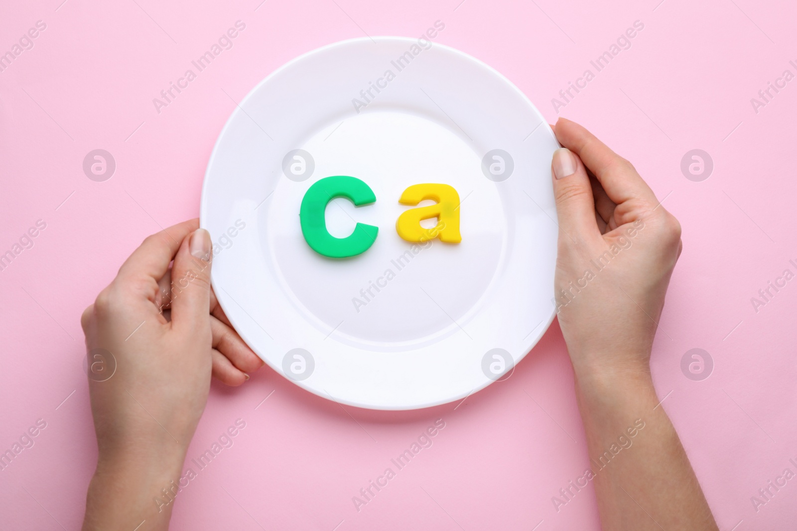 Photo of Woman holding plate with calcium symbol made of colorful letters on pink background, top view