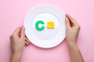 Photo of Woman holding plate with calcium symbol made of colorful letters on pink background, top view