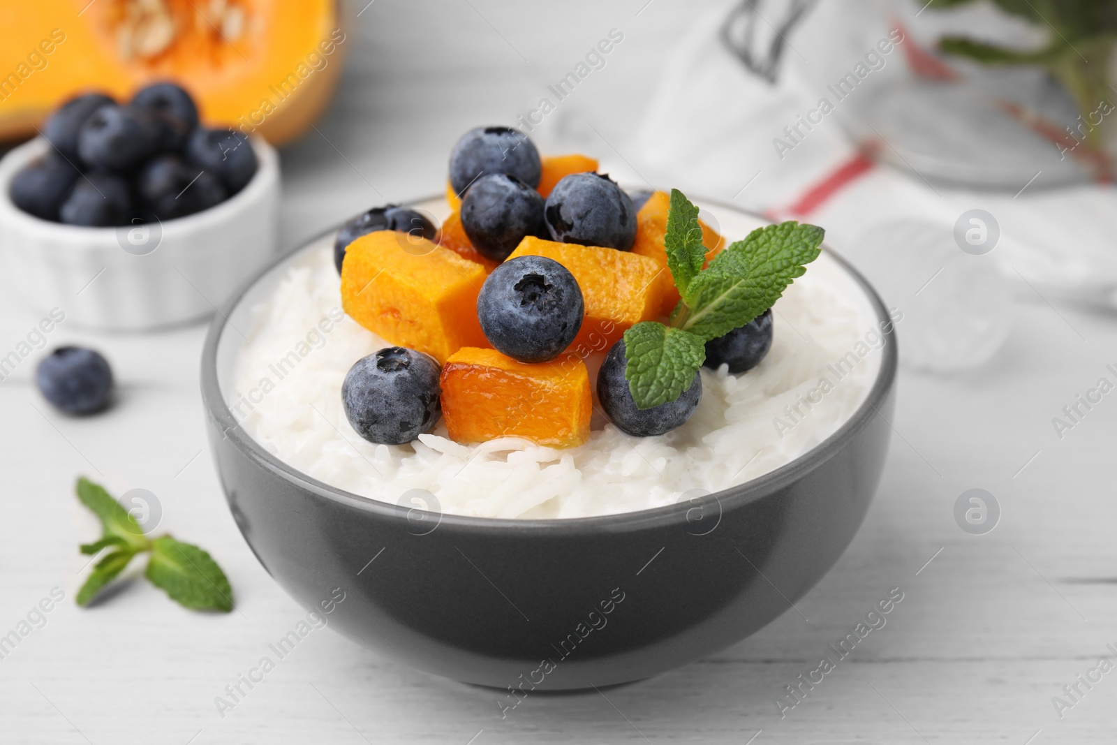 Photo of Bowl of delicious rice porridge with blueberries, pumpkin and mint on white table, closeup