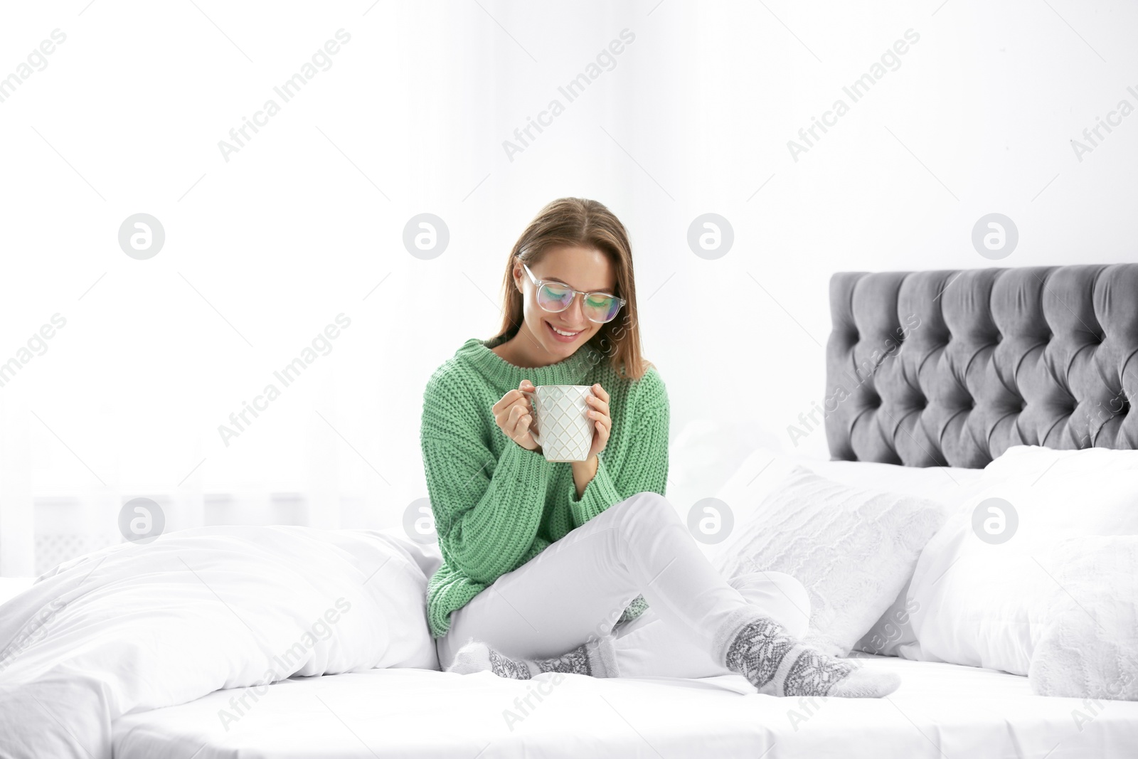 Photo of Young woman in warm sweater with hot drink on bed at home