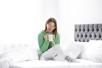 Young woman in warm sweater with hot drink on bed at home