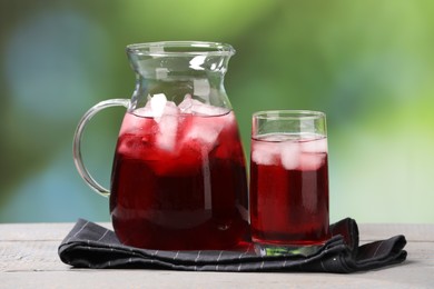 Photo of Refreshing hibiscus tea with ice cubes on white wooden table against blurred green background