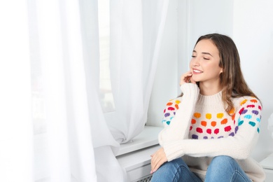 Beautiful teenage girl in warm cozy sweater looking out window at home. Space for text