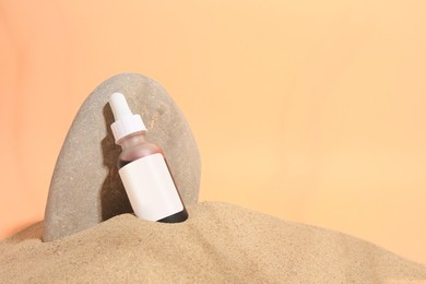 Photo of Bottle with serum and stone on sand against orange background, space for text. Cosmetic product