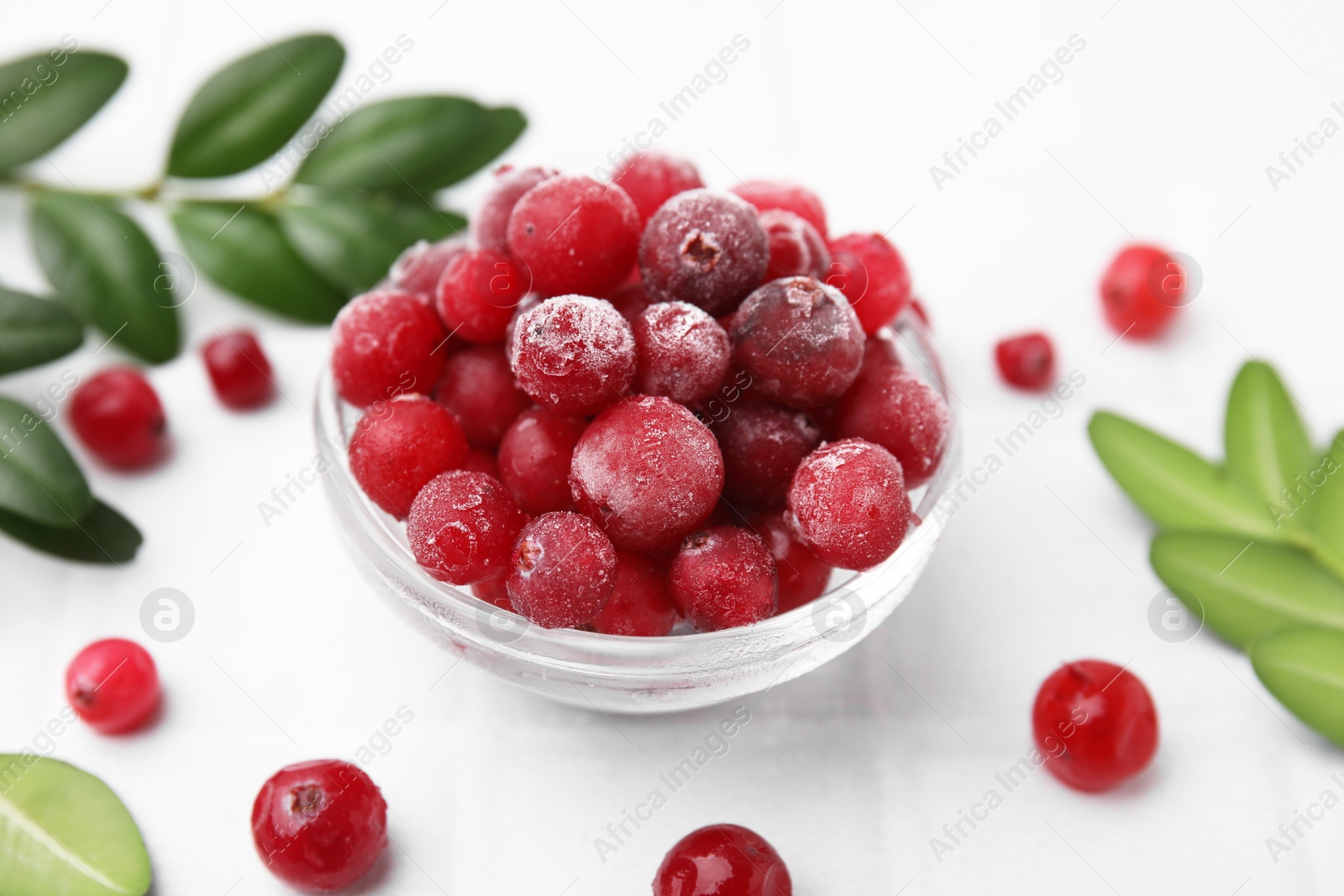 Photo of Frozen red cranberries in bowl and green leaves on white tiled table, closeup