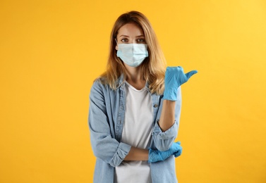 Photo of Young woman in medical gloves and protective mask pointing thumb aside on yellow background