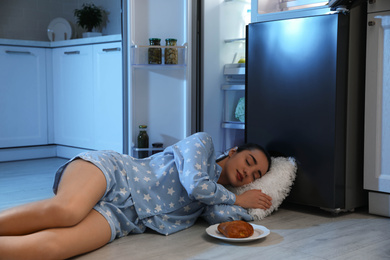 Photo of Young woman sleeping near open refrigerator at night