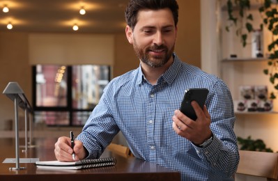 Photo of Handsome man with smartphone writing something in notebook at table in cafe