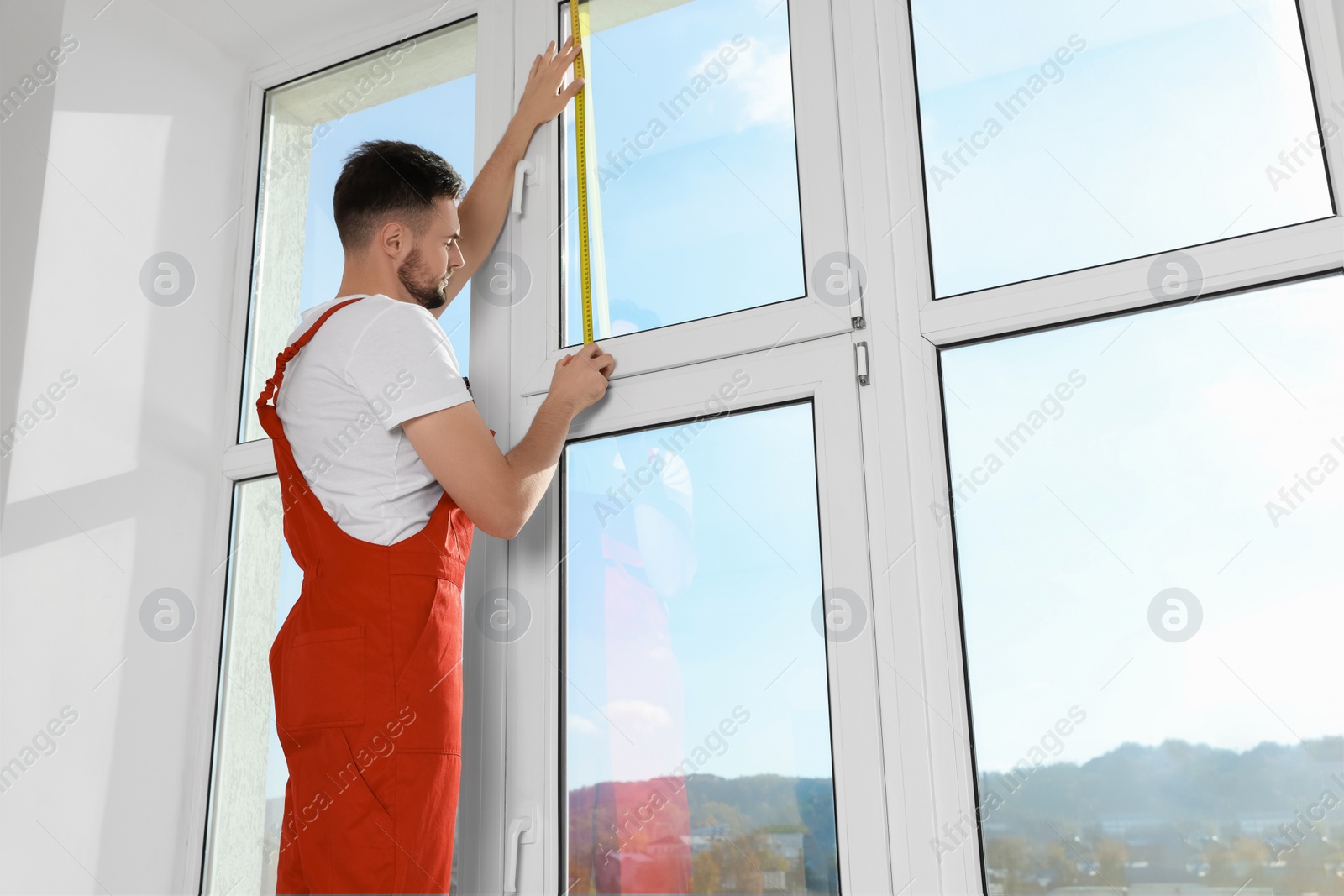 Photo of Worker in uniform measuring window with tape indoors. Roller blinds installation