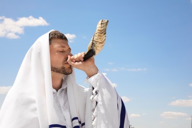 Photo of Jewish man in tallit blowing shofar outdoors. Rosh Hashanah celebration