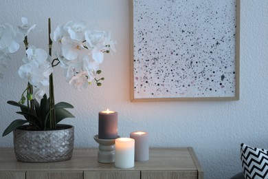 Photo of Beautiful orchid flowers and burning candles on wooden chest of drawers indoors