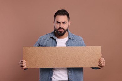 Angry young man holding blank cardboard banner on brown background, space for text
