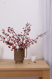 Photo of Hawthorn branches with red berries and candle on wooden table indoors