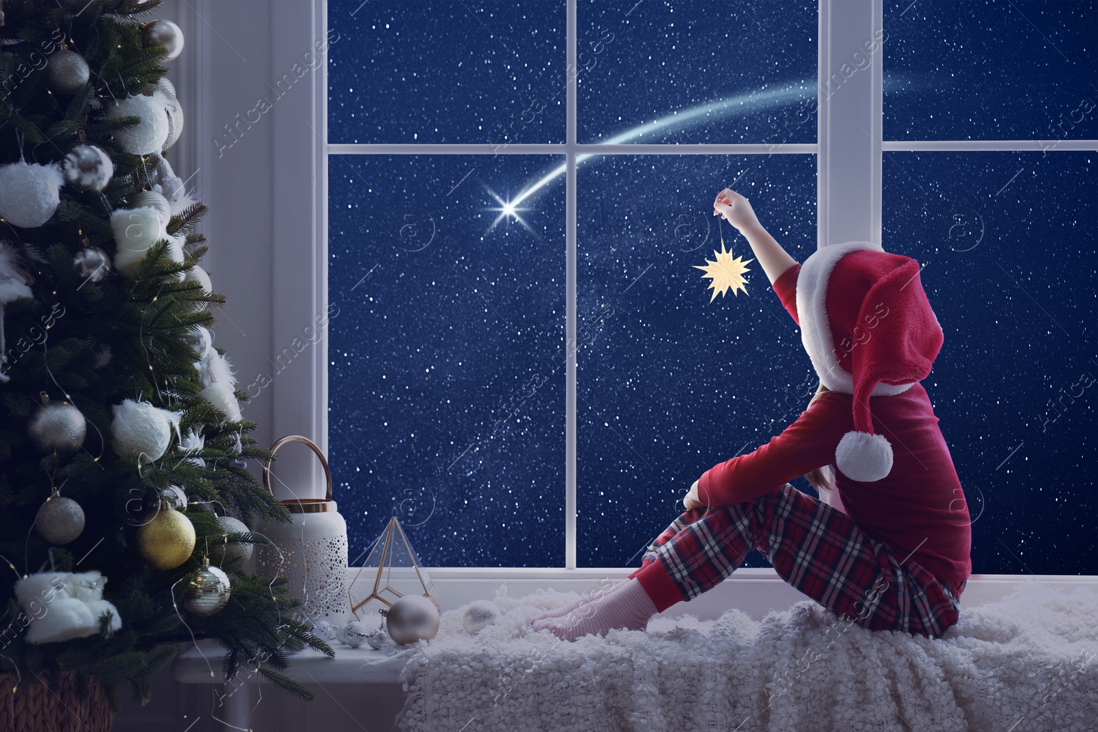 Image of Cute little girl in Santa hat with Christmas ornament sitting on windowsill and looking at shooting star in beautiful night sky