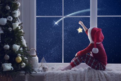 Image of Cute little girl in Santa hat with Christmas ornament sitting on windowsill and looking at shooting star in beautiful night sky