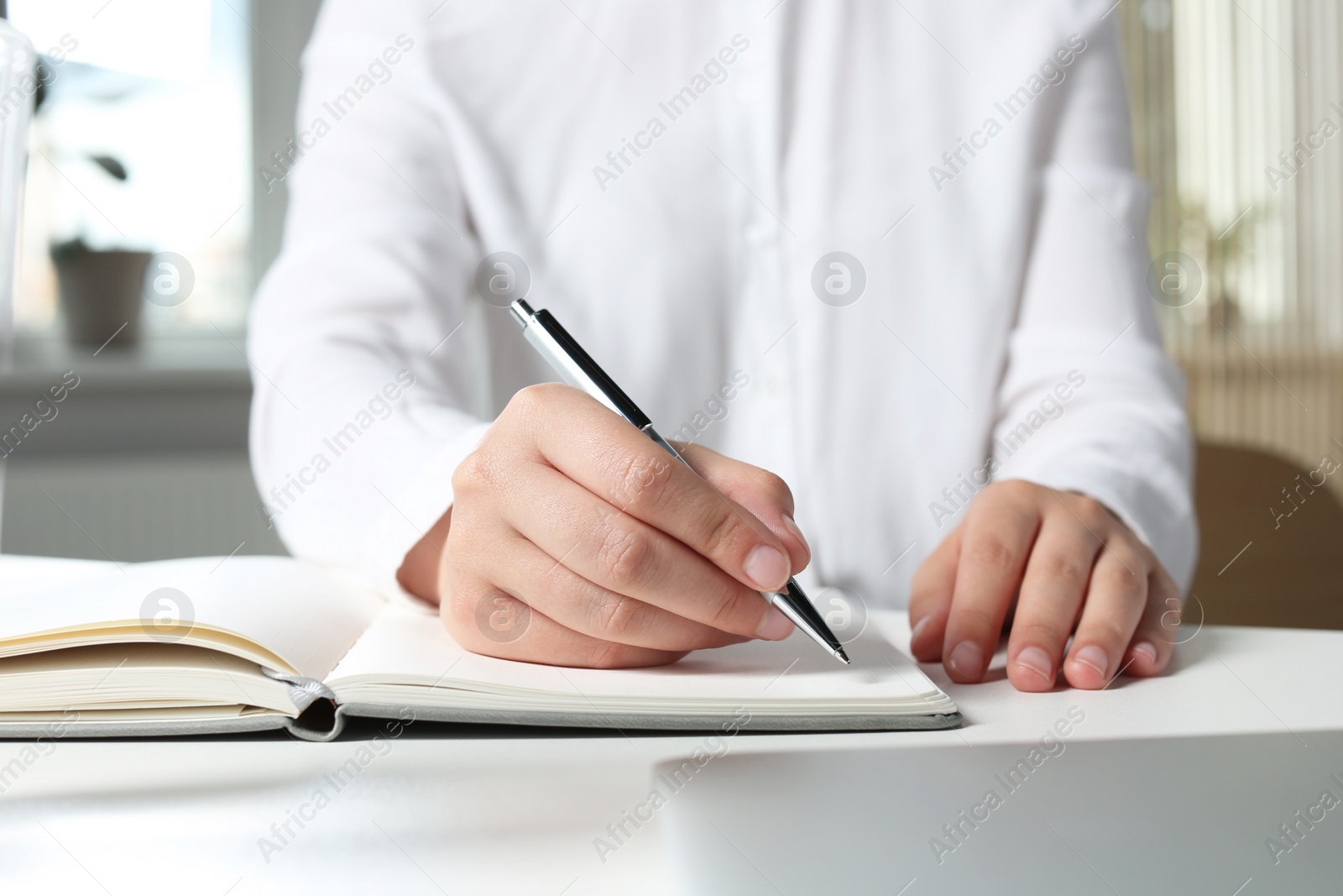 Photo of Woman writing in notebook at white table in office, closeup