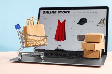 Photo of Online store. Laptop, mini shopping cart and purchases on beige table