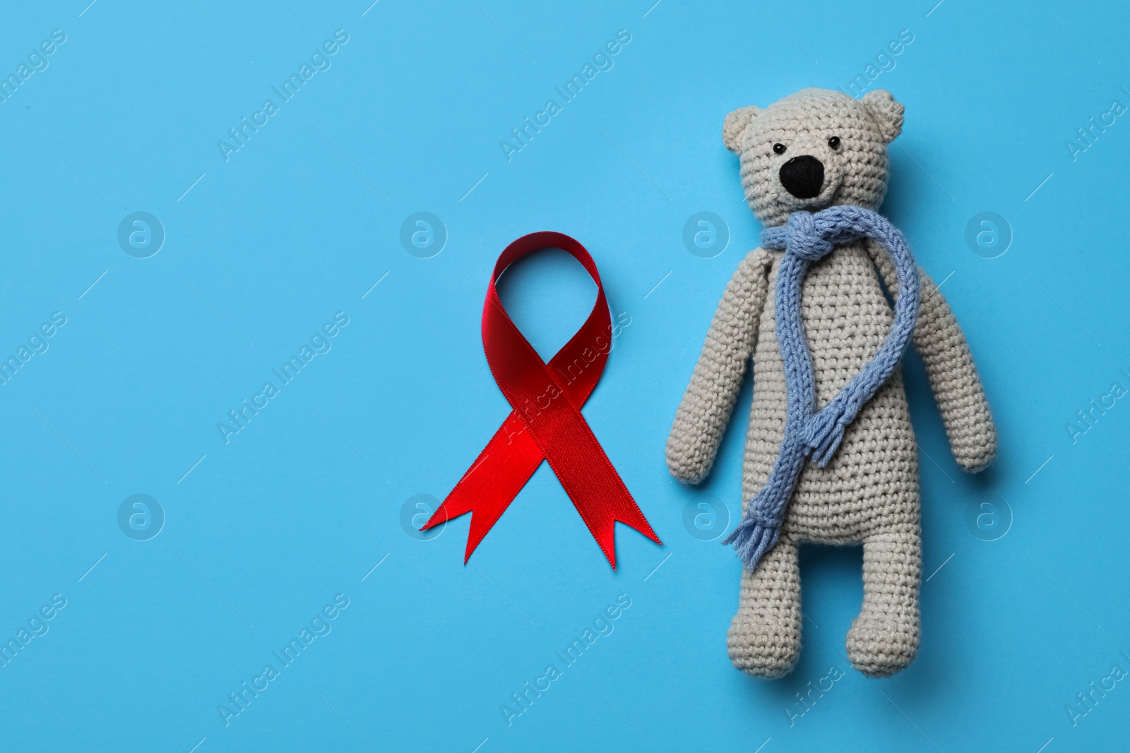 Photo of Cute knitted toy bear and red ribbon on blue background, flat lay with space for text. AIDS disease awareness