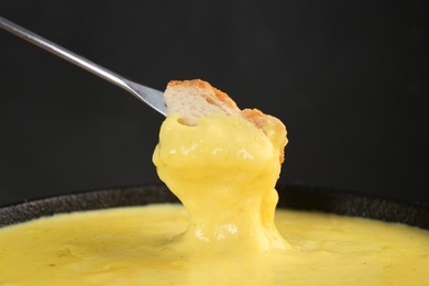 Photo of Dipping piece of bread into fondue pot with tasty melted cheese against dark gray background, closeup