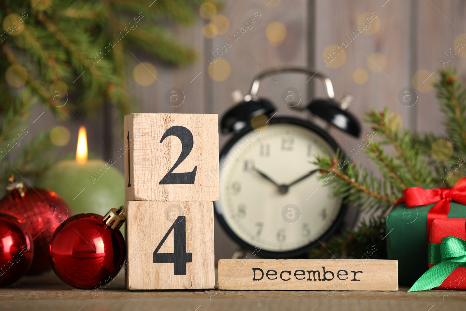Photo of December 24 - Christmas Eve. Wooden block calendar, alarm clock and festive decor on table against blurred lights