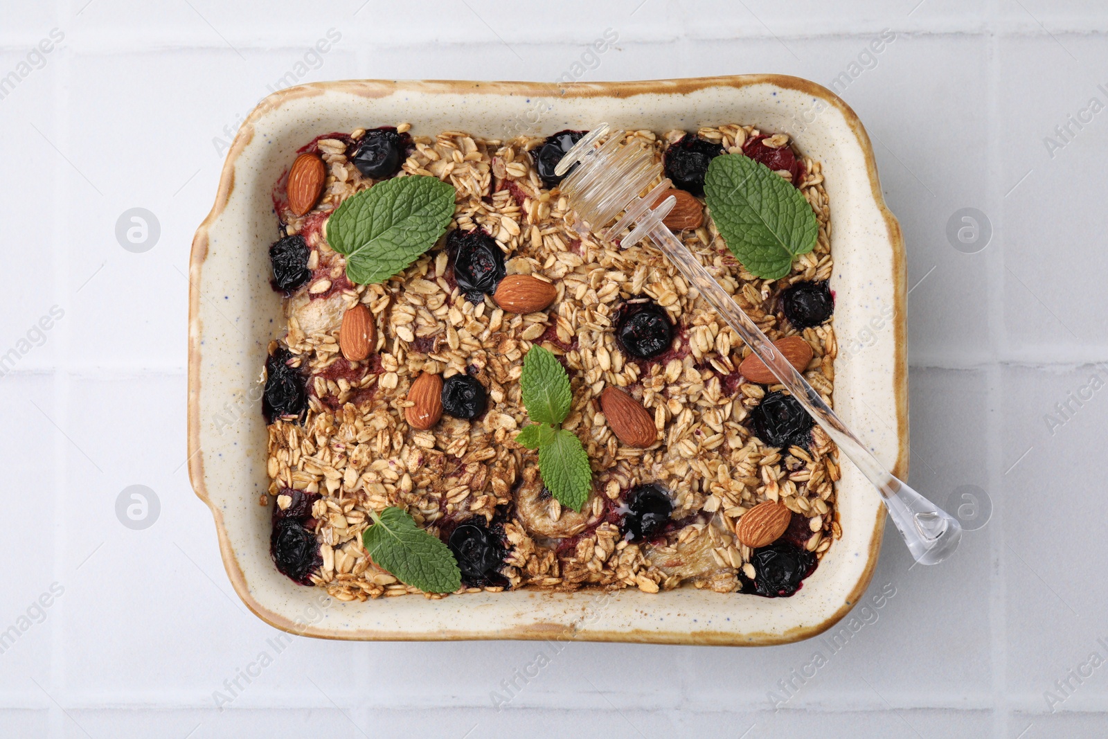 Photo of Tasty baked oatmeal with berries, almonds and honey dipper in baking tray on white tiled table, top view