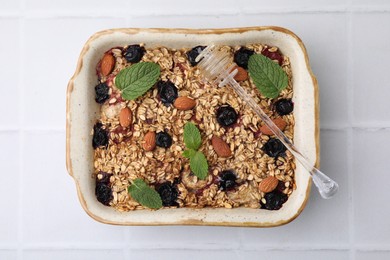 Photo of Tasty baked oatmeal with berries, almonds and honey dipper in baking tray on white tiled table, top view